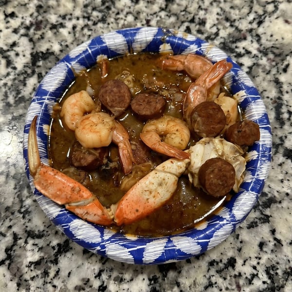 Seafood gumbo with shrimp, sausage, and crab claws in a blue and white paper bowl.