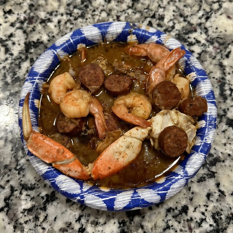Seafood gumbo with shrimp, sausage, and crab claws in a blue and white paper bowl.