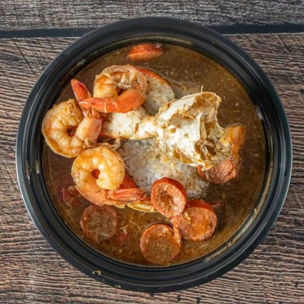 A black bowl of gumbo with shrimp, crab, sausage, and rice on a wooden table.