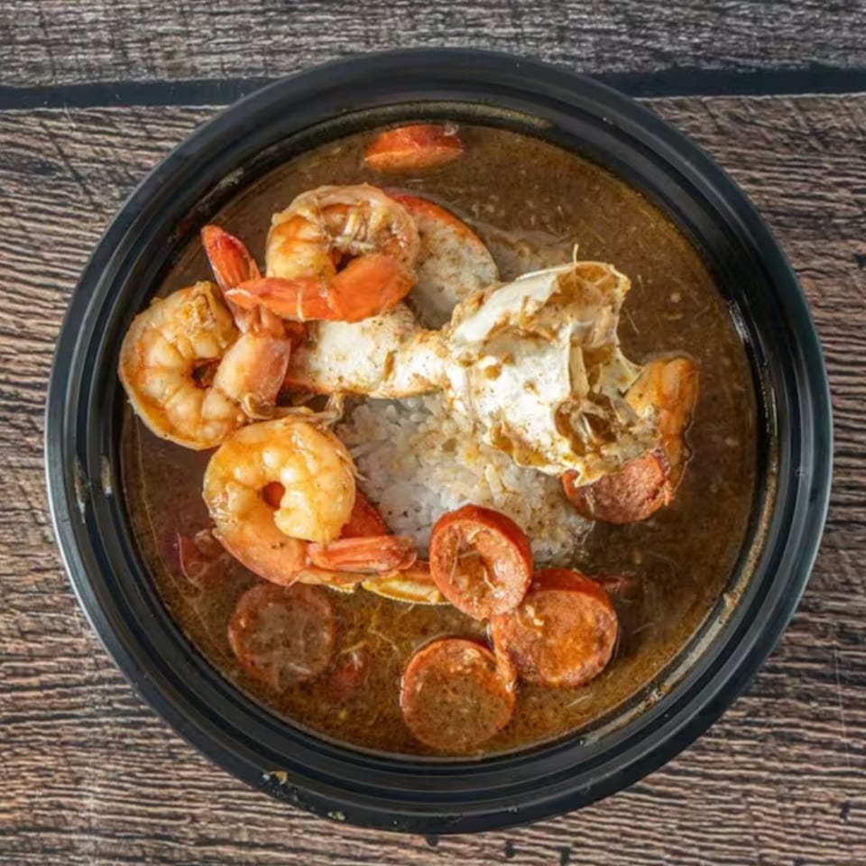 A black bowl of gumbo with shrimp, crab, sausage, and rice on a wooden table.